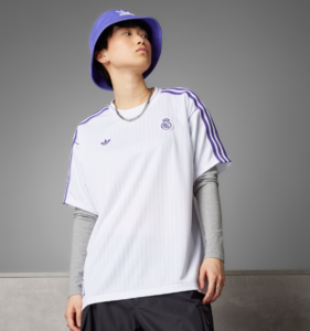 Model wearing a white Real Madrid jersey with purple accents, layered gray sleeves, and a purple bucket hat against a gray backdrop.