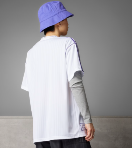 Person seen from behind wearing a white sports jersey with purple piping and a purple bucket hat, gray long-sleeve layer underneath, standing against a gray backdrop