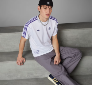 Young man sitting on gray steps in a white Real Madrid jersey with purple stripes, black beanie, and chunky chain necklace.
