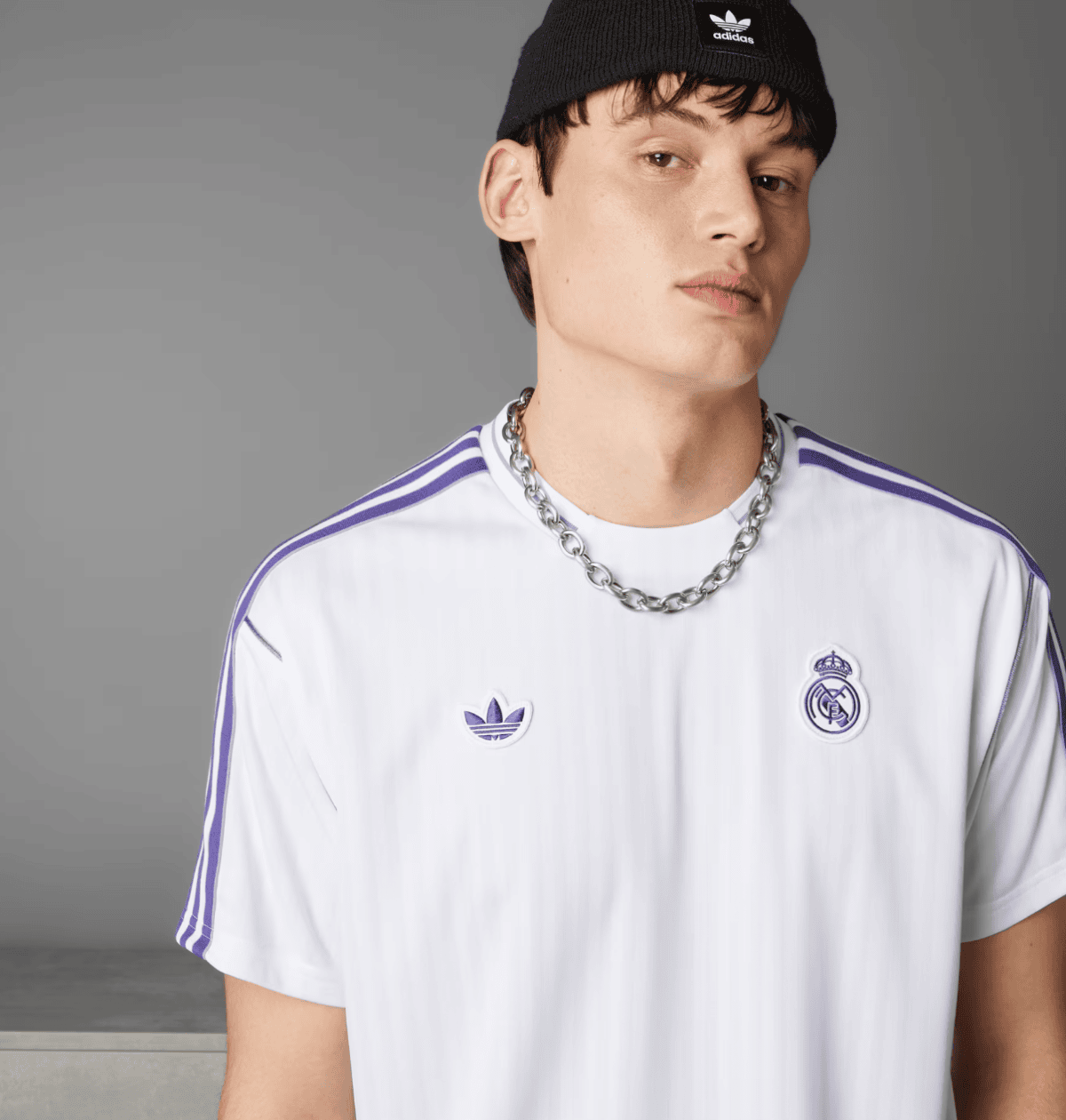 Portrait of a young man wearing a white Real Madrid jersey with purple stripes and a chunky silver chain, with a black beanie, against a gray backdrop.