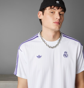 Portrait of a young man wearing a white Real Madrid jersey with purple stripes and a chunky silver chain, with a black beanie, against a gray backdrop.