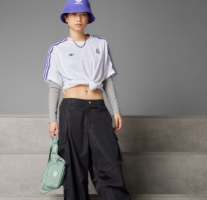 Model wearing a knotted white Real Madrid tee over gray-long sleeves, black cargo pants, purple bucket hat, and carrying a mint green bag.