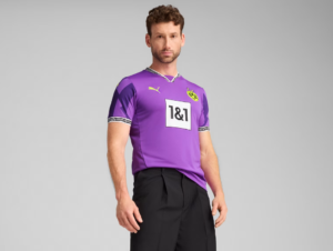 Man posing in a purple Borussia Dortmund football jersey (sponsor 1&1) and black shorts against a gray backdrop.