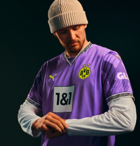 Portrait of a man in a purple Borussia Dortmund jersey and beige knit beanie, looking down and adjusting his white undershirt sleeves.
