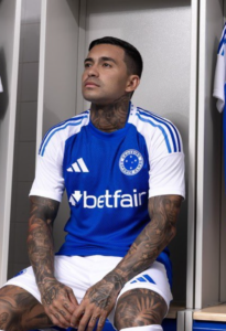 Soccer player with tattoos sits in a locker room wearing a blue and white jersey with a sponsor logo on the chest.