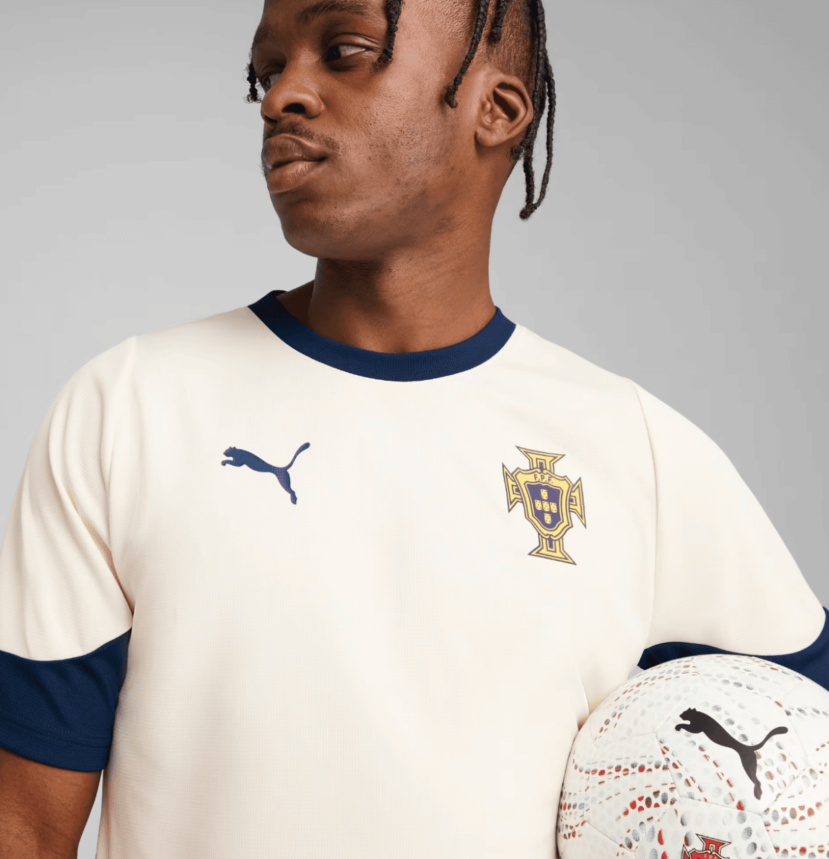 Close-up of a male soccer player in a cream Portugal jersey with navy trim, holding a white football with a blue Puma logo to the right.