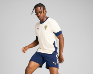 Male model wearing a cream soccer jersey with blue collar and cuffs, crest on the chest, paired with navy shorts with a yellow Puma logo.