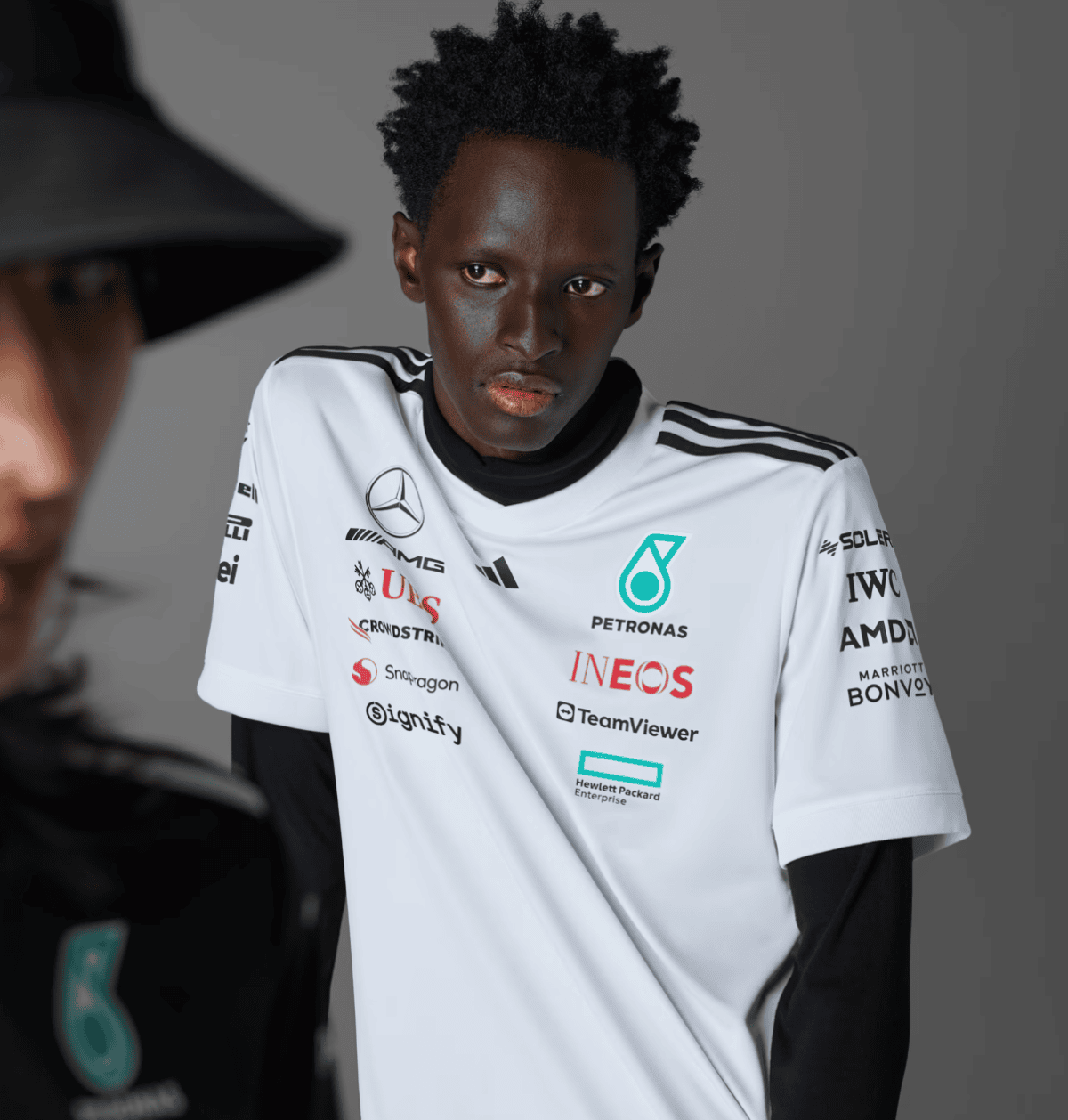 Portrait of a young Black male athlete wearing a white racing jersey with sponsor logos (Petronas, INEOS, TeamViewer) against a gray studio backdrop, looking to the right.