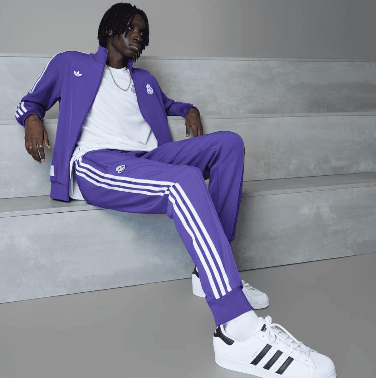 Person wearing a purple Adidas tracksuit with white stripes, white T‑shirt, and white sneakers, sitting on gray concrete steps.