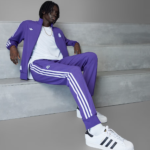 Person wearing a purple Adidas tracksuit with white stripes, white T‑shirt, and white sneakers, sitting on gray concrete steps.