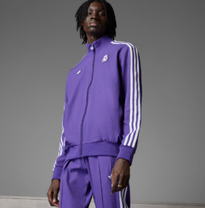 Person wearing a purple Adidas track jacket with white stripes on the sleeves and matching pants, standing against a gray background, model pose.