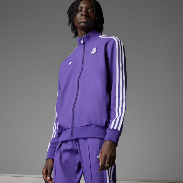 Person wearing a purple Adidas track jacket with white stripes on the sleeves and matching pants, standing against a gray background, model pose.