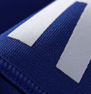 Close-up of blue textile with a white stitched patch forming part of a letter, likely on a sports jersey