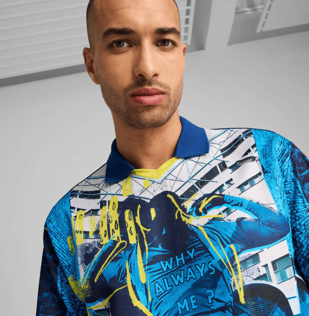 Man with a shaved head wearing a bold, blue and yellow graphic polo shirt, standing against a neutral indoor background.