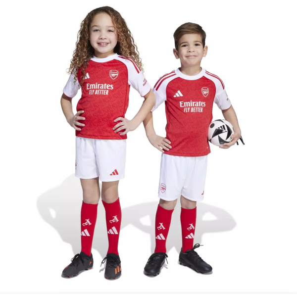 Two smiling children in Arsenal youth soccer kits pose with a ball between them, red jerseys and white shorts.