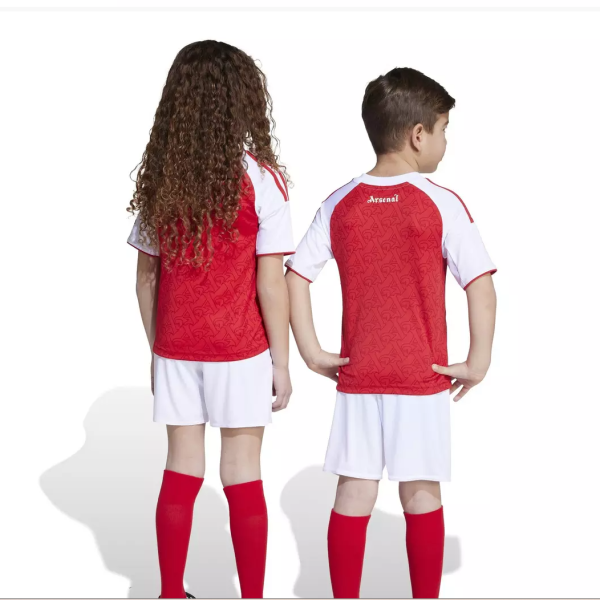 Two children in red and white soccer uniforms stand back to back, facing away from the camera with red socks on the ground side by side.