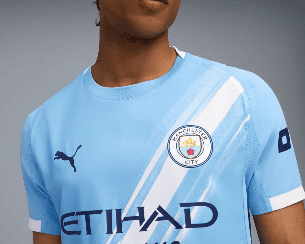 Close-up of a light-blue Manchester City jersey with diagonal white stripes, featuring the club badge and the Etihad sponsor, worn by a person.