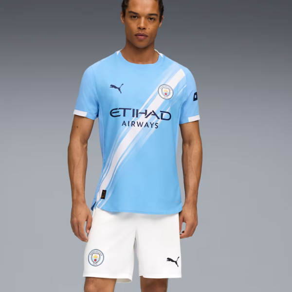 Man wearing Manchester City home kit: light blue jersey with diagonal white stripe and white shorts, crest visible.