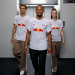 Three athletes in white Red Bull jerseys walk toward the camera in a modern hallway, with an open elevator behind them.