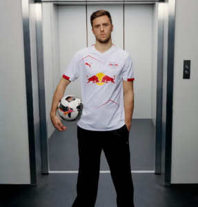 Soccer player in a white Red Bull jersey stands in an elevator, holding a soccer ball at his side and looking at the camera.