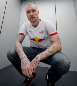 Crouching man with a shaved head and tattoos in a white RB Leipzig jersey, standing in an elevator and looking at the camera.