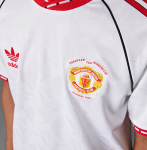 White soccer jersey displaying Adidas logo on the left and Manchester United crest on the right, with red trim along the collar and sleeves.