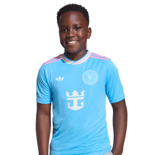 Smiling boy in a light blue soccer jersey with a white crest on the chest, standing against a white background.