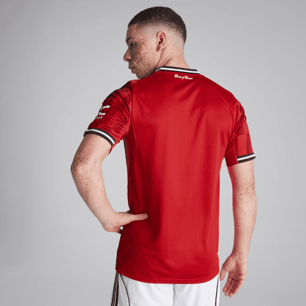 Rear view of a man in a red sports jersey with black-and-white trim, standing with one hand on his hip against a grey studio backdrop.