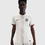 Male model in a white Chelsea FC soccer jersey with a green Nike swoosh and vertical red/black stripes.