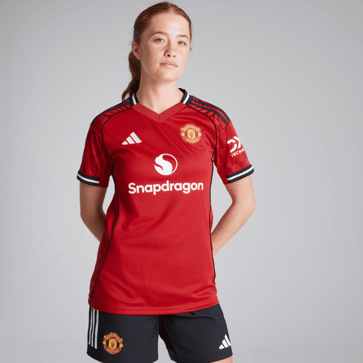 Female soccer player in Manchester United red jersey standing with hands behind back against a gray backdrop, sponsor logos visible (Snapdragon).