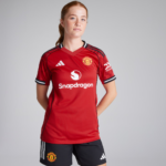 Female soccer player in Manchester United red jersey standing with hands behind back against a gray backdrop, sponsor logos visible (Snapdragon).