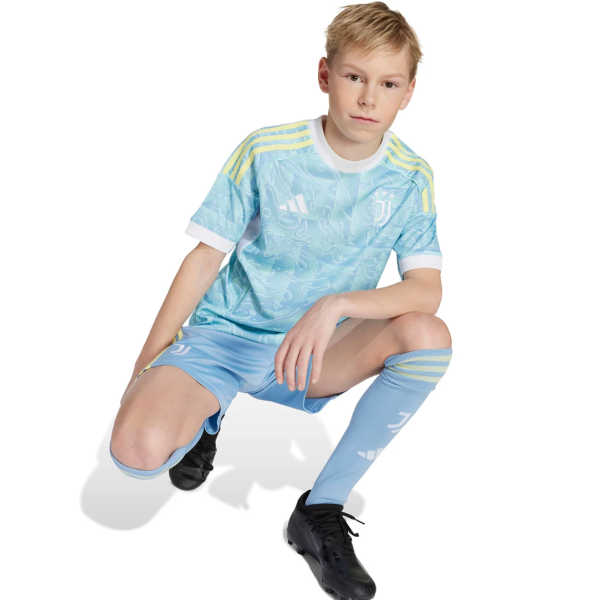 Young boy in a light blue soccer jersey and shorts kneels in a white studio background, wearing matching socks and black cleats.