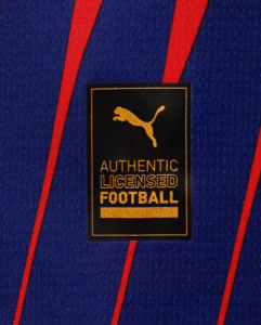 Gold Puma badge on a black patch reading 'AUTHENTIC LICENSED FOOTBALL' sewn onto a blue jersey with red stripes.