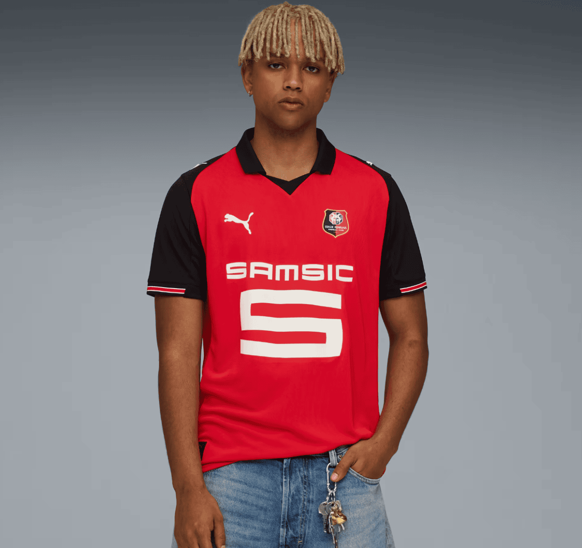 Person standing against a gray gradient background wearing a red and black soccer jersey with a large 'SAMSIC' logo and a team crest on the chest (Puma logo visible).