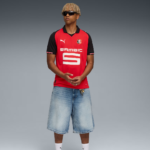 Young man in a red and black soccer jersey with sponsor 'SAMSIC' and crest, wearing sunglasses, standing with hands clasped in front against a gray gradient background.