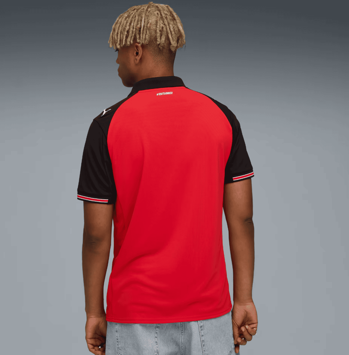 Back view of a person wearing a red polo-style shirt with black sleeves and white-red trim, standing against a gray gradient background, with blond dreadlocks visible
