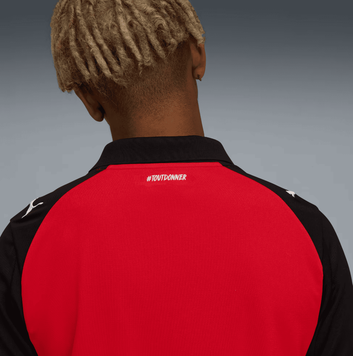 Back view of a person in a red and black athletic jersey; blonde dreadlocks show above the collar, with '#TOUTDONNER' on the upper back.