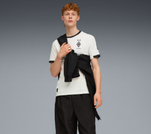Male model wearing a white soccer jersey with crest, black shorts, and a black jacket draped over his shoulder against a gray gradient backdrop.