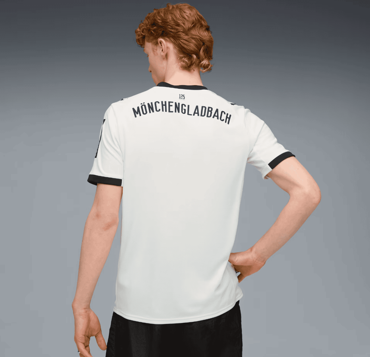 Back view of a person wearing a white soccer jersey with 'MÖNCHENGLADBACH' printed across the top, black collar and sleeve trim, against a gray gradient background.