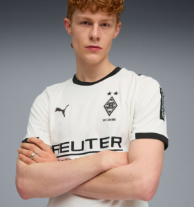 Portrait of a young man with red hair and a serious expression, arms crossed, wearing a white Borussia Mönchengladbach jersey against a gray gradient backdrop.