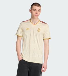 Male model wearing a cream Adidas soccer jersey with gold trim and crest, posing against a light gray background.
