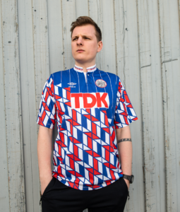 Portrait of a young man wearing a blue, red, and white geometric soccer jersey with 'TDK' logo, standing against a grey corrugated metal wall, hands in pockets.
