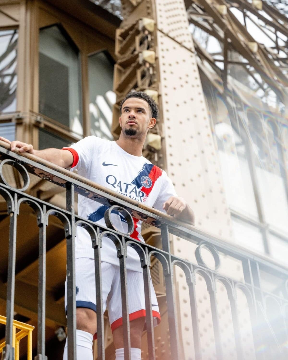 Athlete wearing a PSG jersey stands at a railing on a balcony, gazing into the distance with a metal bridge structure behind him.