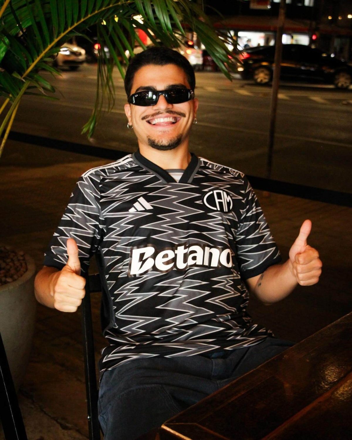 Smiling man wearing sunglasses and a black zigzag-pattern jersey with Betano logo gives thumbs up on a nighttime city street.
