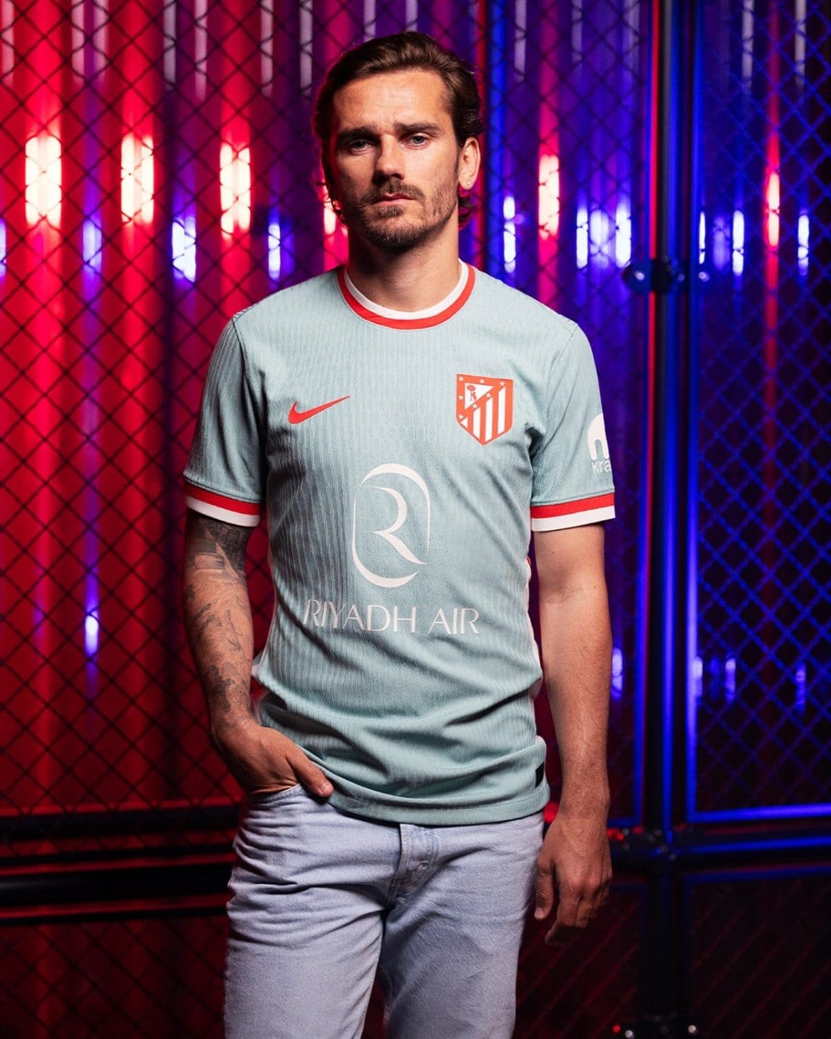 Portrait of a man in a light blue football jersey with red trim, standing in a neon-lit red and blue backdrop behind a chain-link fence.