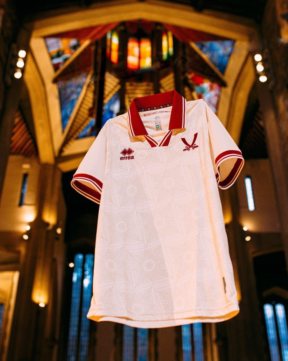 White polo shirt on a display stand with red collar trim, Errea logo on the chest, showcased in a warm, vaulted interior.