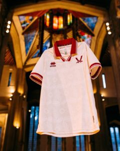 White polo shirt on a display stand with red collar trim, Errea logo on the chest, showcased in a warm, vaulted interior.