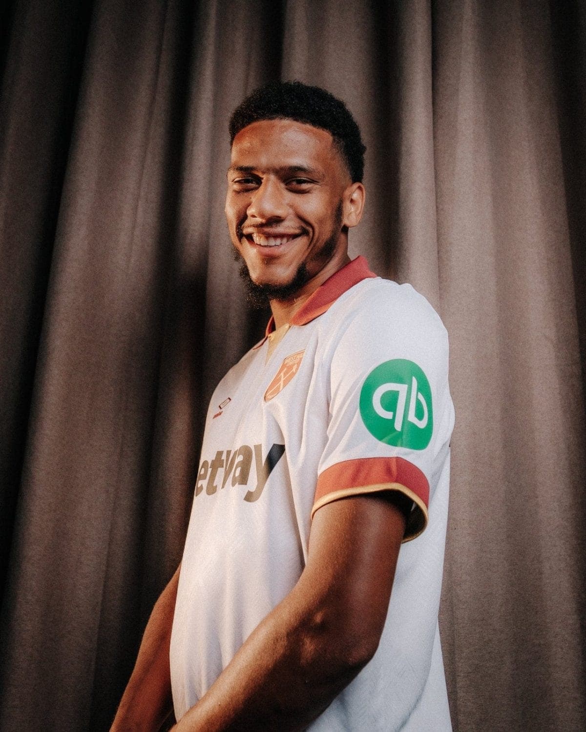Smiling young man in a white team jersey with red trim, standing in front of dark stage curtains.