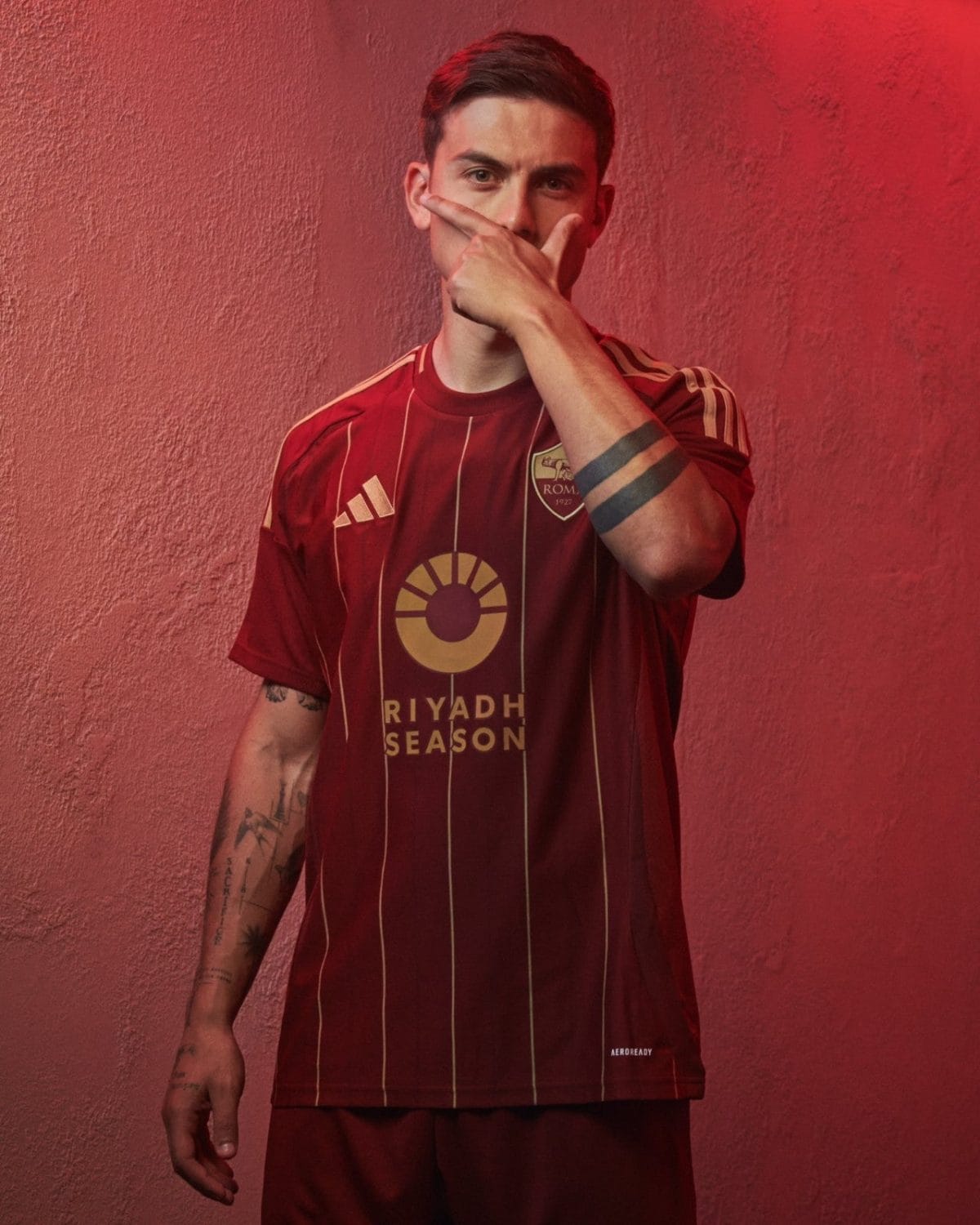Young man in a maroon Riyadh Season football jersey stands against a red textured wall, hand covering his mouth with a partial glare visible behind his fingers.