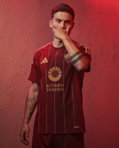 Young man in a maroon Riyadh Season football jersey stands against a red textured wall, hand covering his mouth with a partial glare visible behind his fingers.